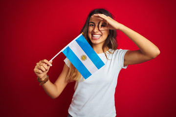 Young beautiful woman holding argentine flag over red isolated background with happy face smiling doing ok sign with hand on eye looking through fingers