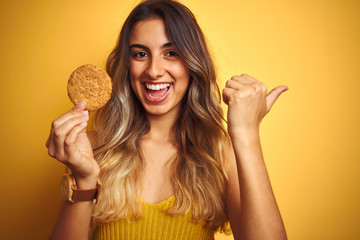 Young beautiful woman eating biscuit over grey isolated background pointing and showing with thumb...