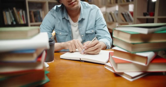 Asian student with curly hair is in library, sits at desk full of books. Guy wearing glasses preparing for exam and studying - education concept close up 4k