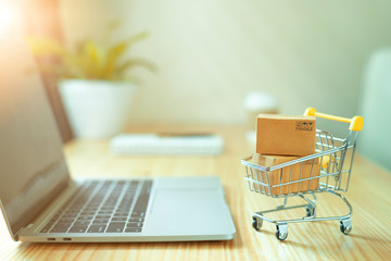 Brown paper boxs in a shopping cart with laptop keyboard on wood table in office background.Easy shopping with finger tips for consumers.Online shopping and delivery service concept. - obrazy, fototapety, plakaty