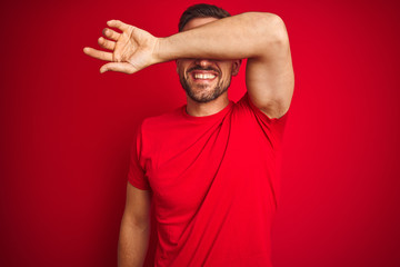 Young handsome man wearing casual t-shirt over red isolated background covering eyes with arm smiling cheerful and funny. Blind concept.