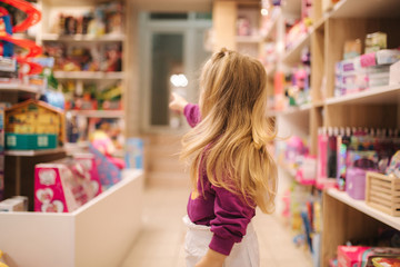 Back view of adorable little girl shopping for toys. Cute female in toy store. Happy young girl...