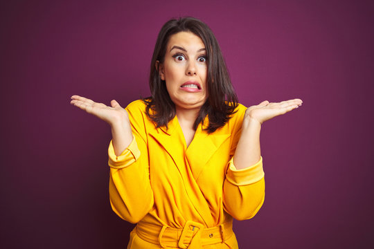 Young beautiful brunette woman wearing elegant yellow jacket over purple isolated background clueless and confused expression with arms and hands raised. Doubt concept.
