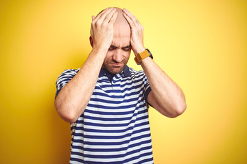 Young bald man with beard wearing casual striped blue t-shirt over yellow isolated background suffering from headache desperate and stressed because pain and migraine. Hands on head.