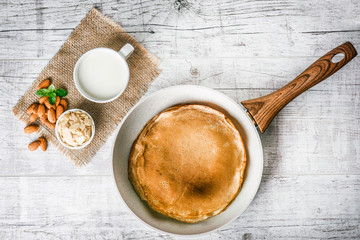 Frying white pancake pan with baked pancake on white rustic table. Almonds and cup of milk on jute and green mint leaf on table. © Milan