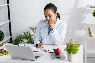 attractive woman in white coat writing in notebook near laptop and vegetables
