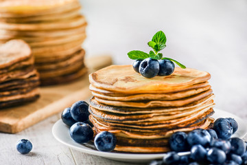 Pancakes with blueberries and mint leaf on top. Pile of small homemade pancakes with forest fruits. Heap of flat thin cake in background.