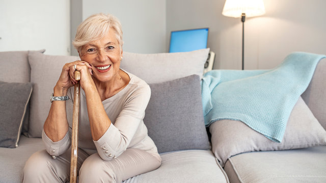 Smiling grandmother sitting on couch. Portrait of a beautiful smiling senior woman with walking cane on light background at home. Old woman sitting with her hands on a cane