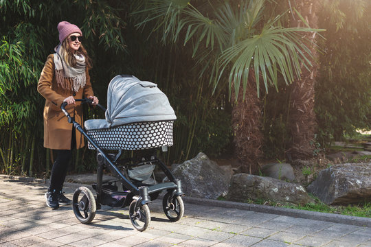 Young Woman In Southern City Or Urban Park Walking Down Street Pulling Stroller. Sunny Spring Or Autumn Weather For Outdoor Activities With Children. Stylish Mother Of Newborn Taking Care Of Herself.