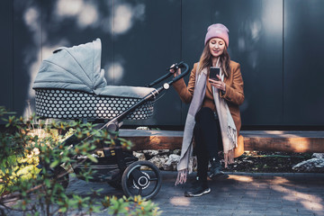 Young mother sitting on bench by fence and communicating on smartphone. Female texting friends...