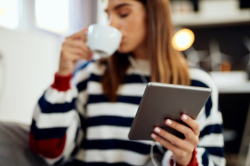 Beautiful caucasian brunette dressed in striped sweater sitting on bed in bedroom, drinking her morning coffee and using tablet.