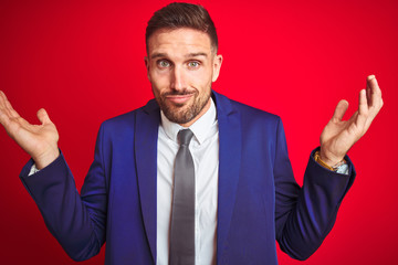 Young handsome business man over red isolated background clueless and confused expression with arms...