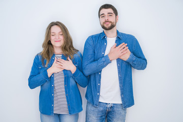 Young beautiful couple standing together over white isolated background smiling with hands on chest with closed eyes and grateful gesture on face. Health concept.