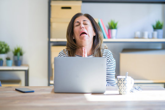 Middle age senior woman sitting at the table at home working using computer laptop begging and praying with hands together with hope expression on face very emotional and worried