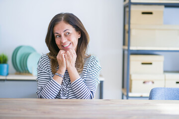 Middle age senior woman sitting at the table at home laughing nervous and excited with hands on...