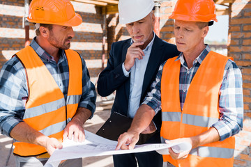 mature constructor pointing with finger at blueprint near businessman