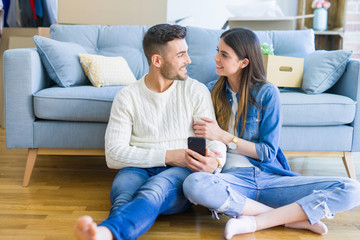 Young couple sitting on the floor of new home using smartphone and smiling happy for moving to a new home