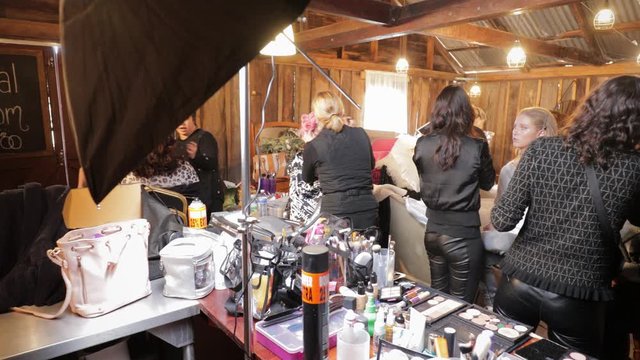 Five beautiful models being prepared for photoshoot by professional hair and make-up artists and stylists in cramped wooden building with tin roof. Empowerment, concentration and focus. 4K