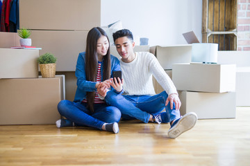 Young asian couple sitting on the floor of new apartment arround cardboard boxes, using smartphone...