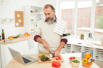 Waist up portrait of bearded senior man watching video recipe via laptop while cooking vegetables in kitchen, copy space