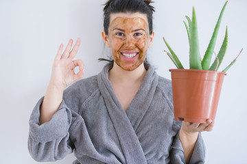 Beautiful woman wearing cosmetic facial mask as skincare beauty treatment and holding aloe doing ok sign with fingers, excellent symbol