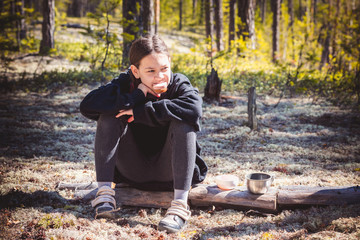 Fototapeta premium An adult girl in warm sweater sits on fallen tree and eats sandwich with sausage resting after hike in pine forest.
