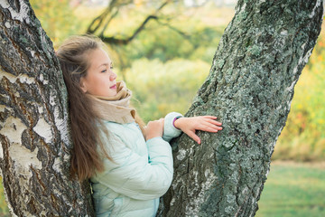 Pensive girl in blue down jacket leaning on birch tree on an autumn day.