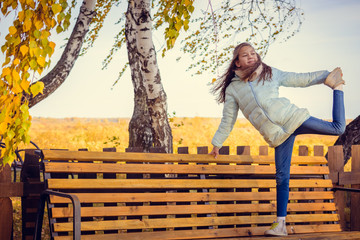 Sports girl in light down jacket doing stretching legs on bench on morning of autumn.