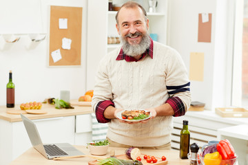 Waist up portrait of bearded senior man presenting meat steak and smiling at camera while cooking in kitchen, copy space