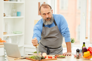 Portrait of tough bearded man holding knife and looking at camera while cooking meat in kitchen, copy space
