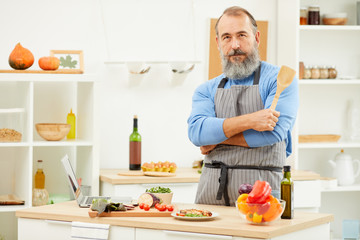 Waist up portrait of bearded senior man cooking at home and looking at camera while posing confidently in kitchen, copy space