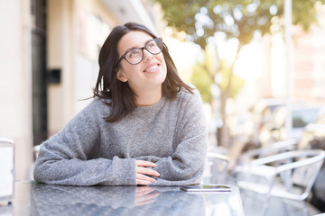 Beautiful young girl wearing glasses smiling cheerful sitting on a cafeteria, relaxing and enjoying sunlight at the town