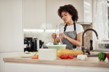 Attractive mixed race woman with curly hair and in apron mixing salad while standing in domestic kitchen.