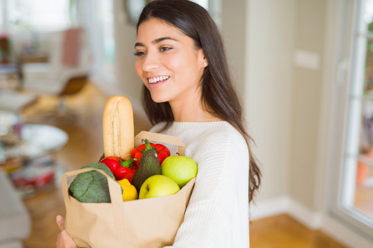 Beautiful young woman smiling holding a paper bag full of fresh groceries at home