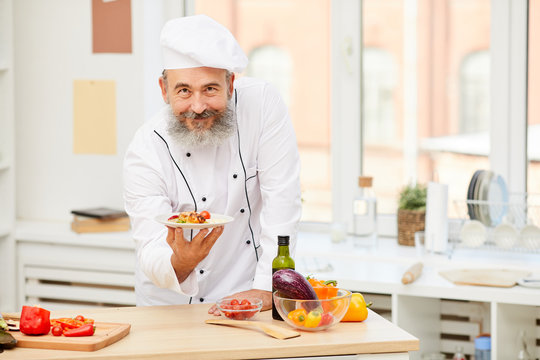 Portrait of cheerful senior chef presenting beautiful dish while posing in restaurant kitchen, copy space