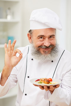 Portrait of cheerful senior chef presenting beautiful Italian dish and showing OK sign while posing in restaurant kitchen