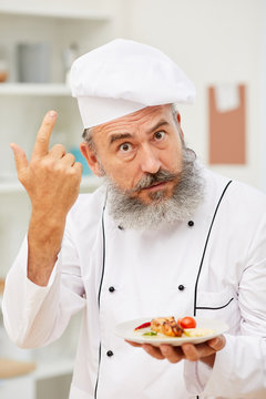 Portrait of cheerful senior chef presenting beautiful Italian dish and looking at camera with curious face expression while posing in restaurant kitchen