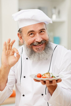 Portrait of bearded senior chef presenting beautiful Italian dish and smiling at camera while posing in restaurant kitchen