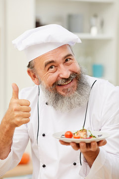 Portrait of cheerful senior chef presenting beautiful Italian dish and showing thumbs up while posing in restaurant kitchen