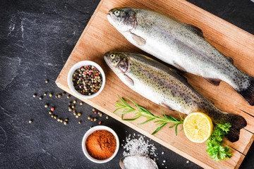 Two whole trouts on wooden board with lemon and rosemary herb. Fishes from top view on dark stone table with pepper and salt..