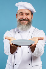 Portrait of bearded senior chef holding empty plate and smiling at camera while standing against blue background, mock up template, focus on foreground