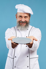 Portrait of bearded senior chef holding empty plate and smiling at camera while standing against blue background, mock up template