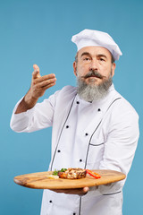 Waist up portrait of charismatic senior chef holding beautiful dish on wooden platter while standing against blue background and looking at camera
