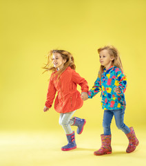 A full length portrait of a bright fashionable girls in a raincoat holding hands, running and having fun on yellow studio background. Autumn and spring fashion for kids. Cute stylish blonde girls.