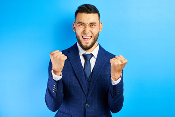 Joyful Asian Kazakh man in business suit and tie clenches fists with happiness and screams, celebrates victory and success isolated on blue wall background