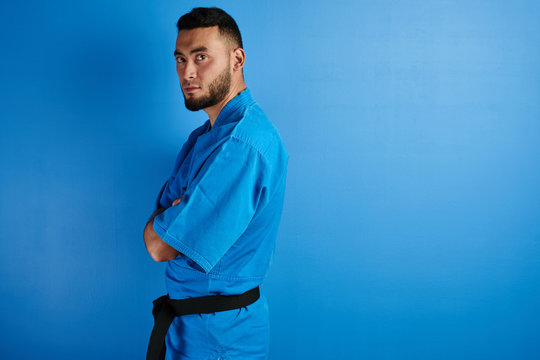 Asian Kazakh karate fighter in kimono uniform stands with arms crossed on blue background with copy space isolated in studio