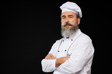 Professional waist up portrait of bearded senior chef posing against black background standing with...