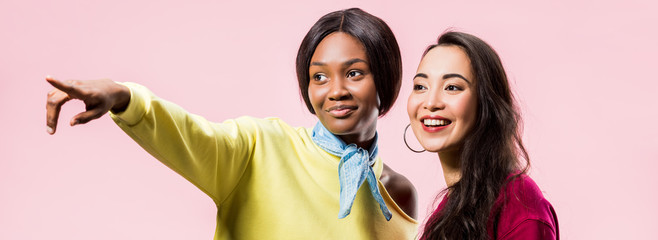 panoramic shot of smiling asian and african american friends pointing with finger isolated on pink