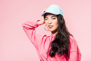 attractive asian woman in pink pullover and cap winking isolated on pink