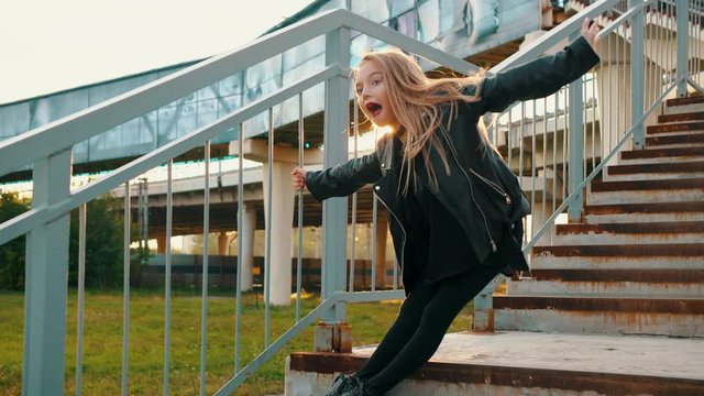 Excited stylish teenage girl posing on stairs during sunset. Low angle view of adorable girl in black leather jacket holding railing and posing on steps during sunset. Fashion concept